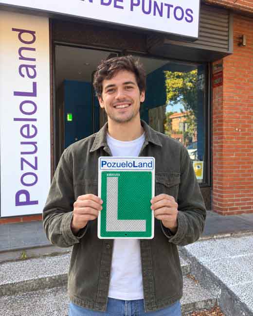 Joven sonriente sosteniendo una placa con la letra L de conductor novel frente a la fachada de una autoescuela en Pozuelo de Alarcón, contento al haber aprobado su carnet tipo B